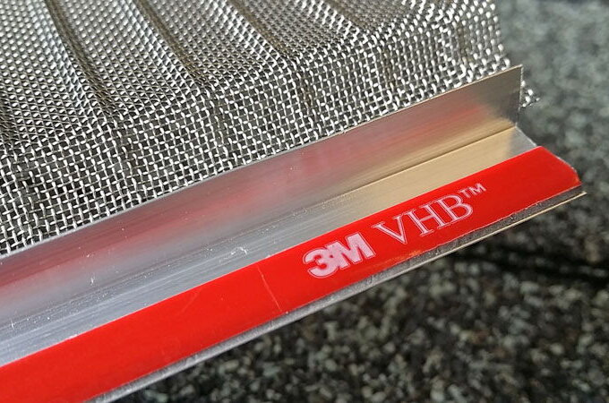 A close-up of a stainless steel micro-mesh screen with an aluminum edge sealed by red 3M VHB™ double-sided tape, displaying the 3M logo and VHB label—similar to products found at Home Depot.