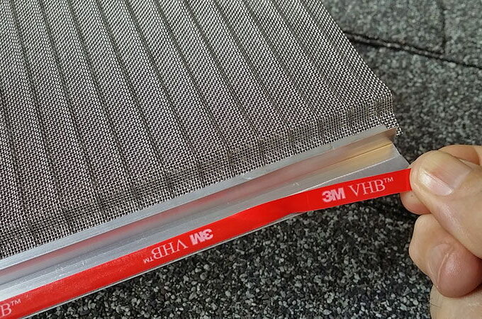 A hand peels back red 3M VHB adhesive tape from the edge of a metal screen frame, revealing the sticky side and the stainless steel micro-mesh of Gutter Guard by Gutterglove, available at Home Depot.