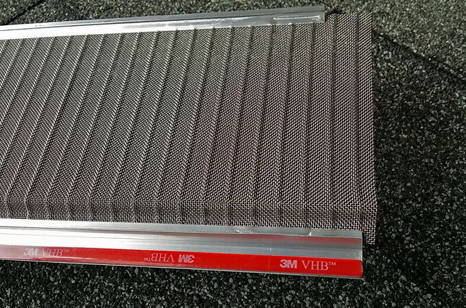 A close-up of a mesh panel with RiserStep Technology, framed with metal strips and resting on a speckled dark surface. The edge features a red adhesive strip labeled 3M VHB™.