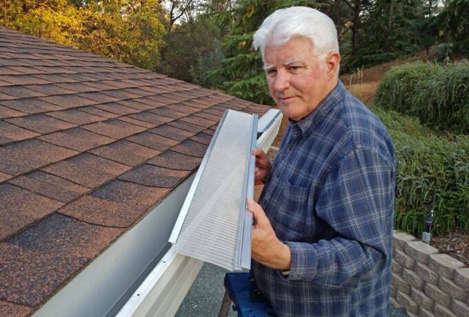 An older man in a plaid shirt stands on a ladder next to a house roof, holding a Gutterglove gutter guard featuring RiserStep Technology above an open rain gutter. Trees and landscaping are visible in the background.