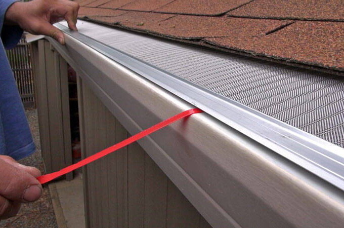 A person installs a stainless steel micro-mesh gutter guard on a roof, using a red tool to ensure proper placement along the gutter’s edge beneath brown roof shingles, similar to options found at Home Depot.