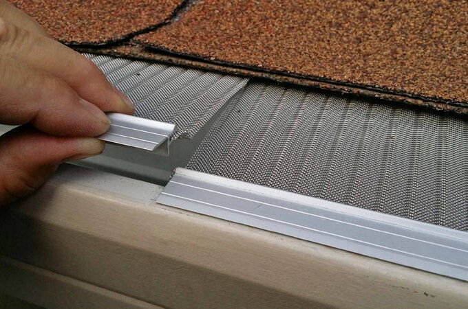 A hand installing a Gutterglove stainless steel micro-mesh gutter guard under roof shingles, demonstrating how it fits over a rain gutter to help prevent debris from entering.