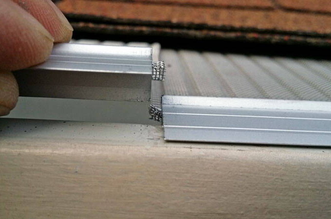 Close-up of fingers holding the edge of a stainless steel micro-mesh frame, aligning it with another metal piece on a flat surface, possibly assembling or repairing a window or door screen.