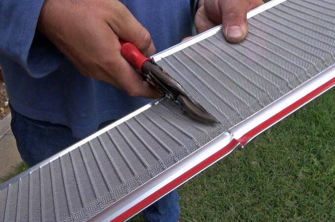 A person uses red-handled tin snips to cut a metal mesh gutter guard with RiserStep Technology, standing outdoors on grass and concrete, similar to products available at Home Depot.