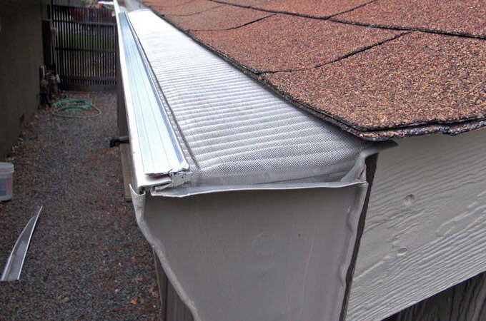 A close-up view of a rooftop gutter protected by a Gutter Guard by Gutterglove, preventing debris from entering, with brown shingles above and gravel ground below.