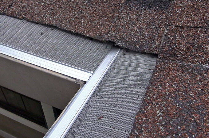 Close-up of a house roof with brown shingles and a Gutter Guard by Gutterglove featuring stainless steel micro-mesh and RiserStep Technology installed along the edge. A window is visible below the roofline.
