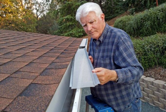 An older man in a plaid shirt stands on a ladder, installing a Gutter Guard by Gutterglove with stainless steel micro-mesh on the roof of a house surrounded by greenery and trees.
