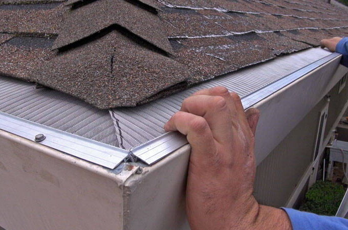 A person installs a stainless steel micro-mesh gutter guard with RiserStep Technology along the edge of a shingle roof to prevent debris from entering the gutter, ideal for Home Depot shoppers.