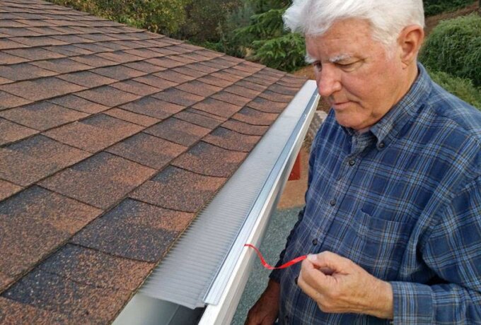 An older man in a plaid shirt stands next to a house roof, inspecting a covered rain gutter with a small red tool near the Gutter Guard by Gutterglove. Trees are visible in the background, suggesting easy installation and same-day pickup from Home Depot.