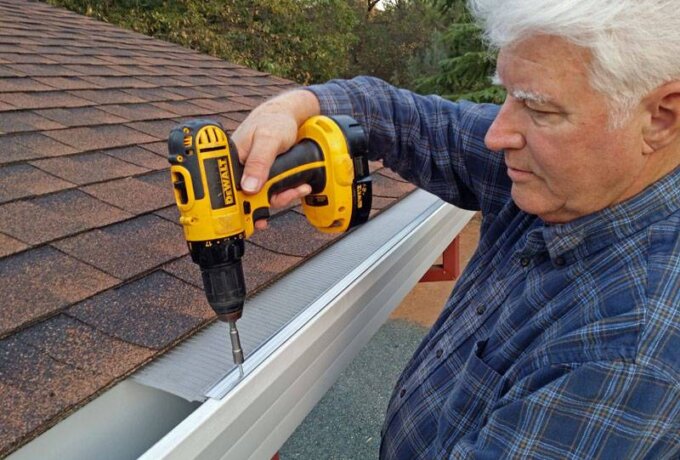 An older man in a blue plaid shirt uses a yellow cordless drill to attach a Gutter Guard by Gutterglove with stainless steel micro-mesh to the edge of a shingled roof. Trees can be seen in the background.
