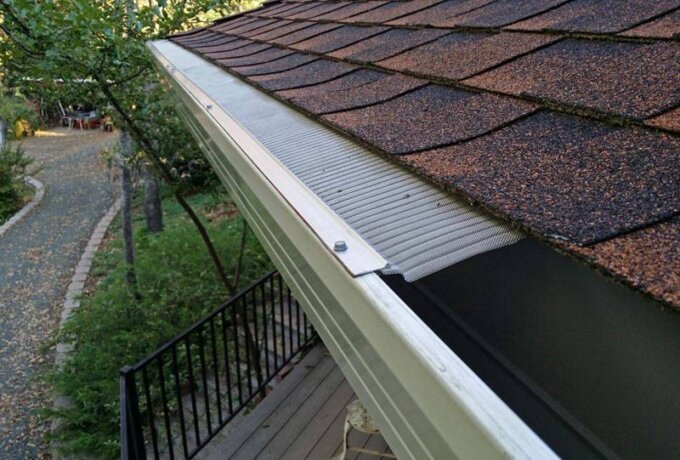 A close-up view of a house roof with brown shingles and a stainless steel micro-mesh Gutter Guard by Gutterglove featuring RiserStep Technology, overlooking a patio and a tree-lined path below.