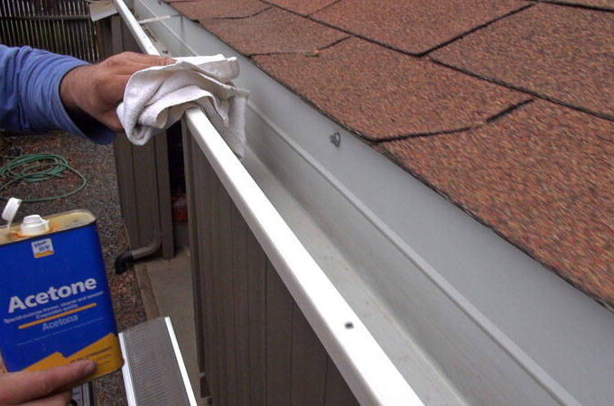 A person wearing a blue sleeve uses a white cloth and a can of acetone to clean the inside edge of a house gutter fitted with Gutter Guard by Gutterglove, beneath brown roof shingles.