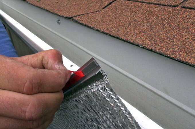 A person installs a stainless steel micro-mesh gutter guard on a roof, securing it along the edge of the shingles to prevent debris from entering the gutter.
