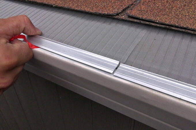 A hand installs a Gutterglove stainless steel micro-mesh gutter guard over a rain gutter, protecting it from debris. The guard features a fine mesh screen and fits under the roof shingles.