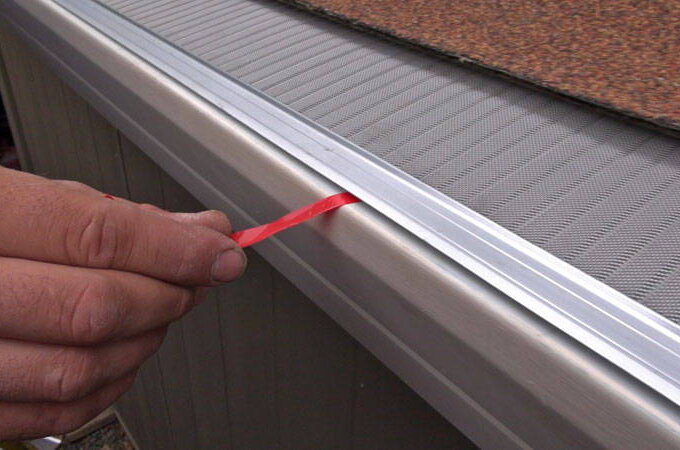 A hand pulls a red plastic strip from the edge of a Gutter Guard by Gutterglove, featuring stainless steel micro-mesh, installed on a house next to a shingled roof.