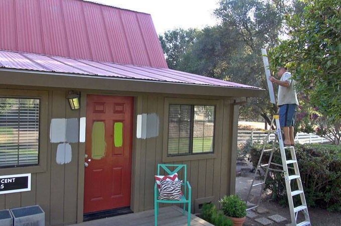 A person stands on a ladder painting the trim of a small house with a red metal roof. The home, featuring Gutter Guard by Gutterglove, has paint samples on the wall and a sign reading Heaven Scent Farms by the door. A chair with cushion sits on the porch.