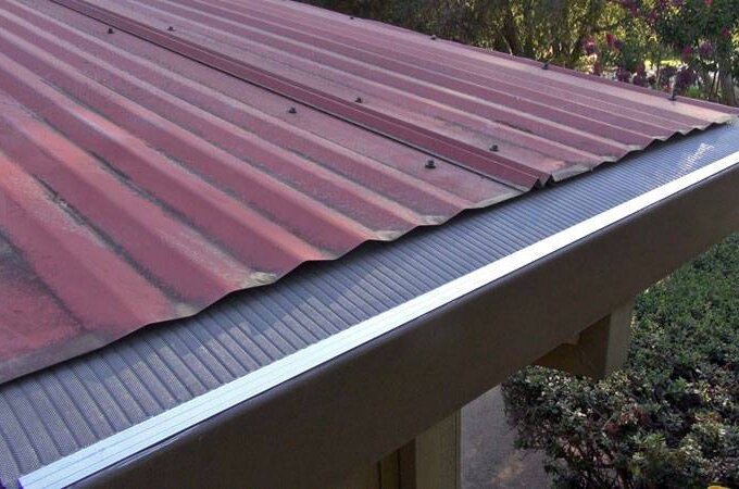 A red metal roof with a Gutter Guard by Gutterglove featuring RiserStep Technology along the edge, next to a garden with green bushes and trees in the background.
