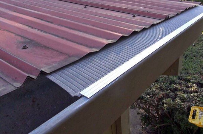 A close-up view of a metal roof with the Gutter Guard by Gutterglove installed along the edge, preventing debris from entering the gutter. Nearby, bushes and a yellow tool are visible below. Perfect for Home Depot shoppers seeking effective protection.