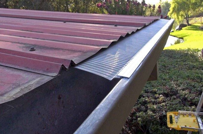 A close-up view of a red metal roof with a stainless steel micro-mesh gutter guard featuring RiserStep Technology installed along the edge, a ladder nearby, and green bushes and trees in the background.