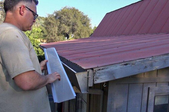 A man stands next to a shed with a red metal roof, holding a long strip of mesh material, appearing to measure or install Gutter Guard by Gutterglove along the roof edge under clear skies. Same-day pickup available at Home Depot.