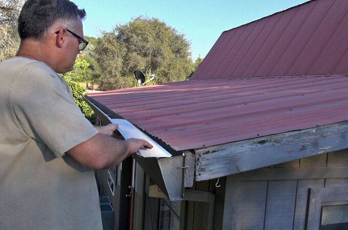 A man wearing glasses installs a metal piece with stainless steel micro-mesh along the edge of a red metal roof on a small wooden building, standing on a ladder with trees visible in the background.