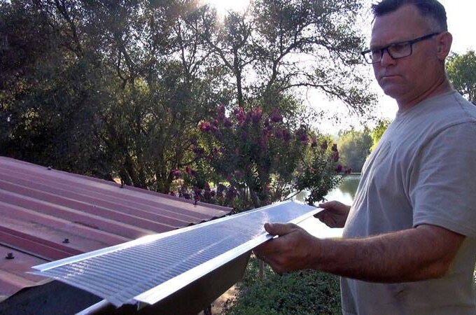 A man wearing glasses holds a Gutterglove metal gutter cover next to a red metal roof, with trees and sunlight in the background of a grassy yard, ideal for those seeking Home Depot same-day pickup.