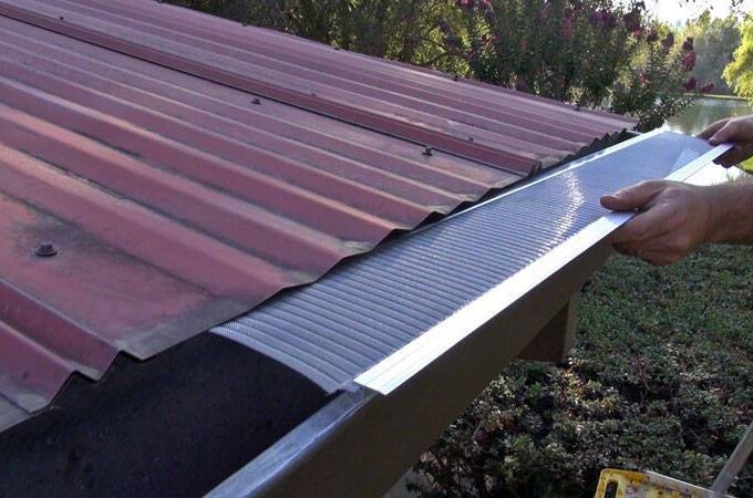 A person installs a metal gutter guard with stainless steel micro-mesh under the edge of a red corrugated metal roof, covering the open gutter to prevent debris from collecting inside. Shrubs and trees are visible in the background.
