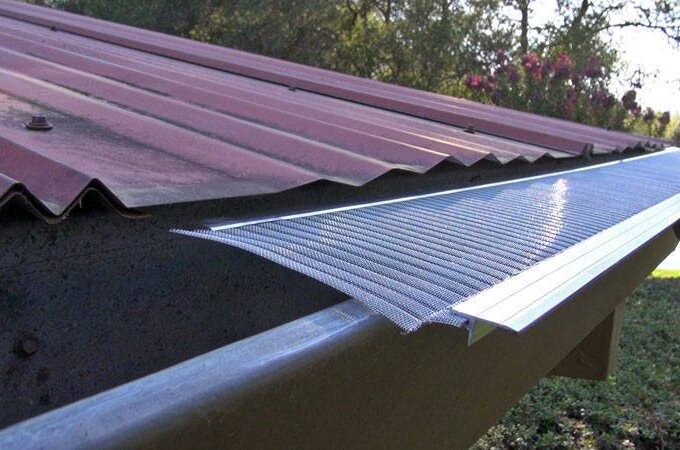 A close-up of a hand installing a stainless steel micro-mesh gutter guard with RiserStep Technology along the edge of a red metal roof, with trees and greenery in the background.