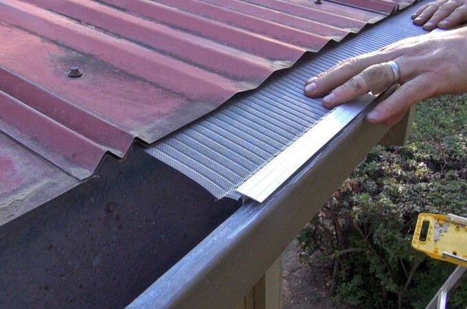 A person installs a Gutter Guard by Gutterglove on the edge of a red corrugated metal roof, pressing the mesh into place above the gutter.