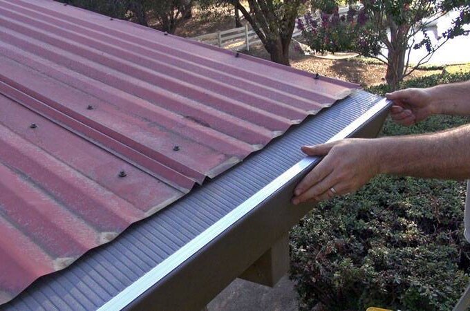 A person installing a Gutterglove gutter guard along the edge of a red metal roof, with trees and bushes in the background; perfect for same-day pickup at Home Depot.