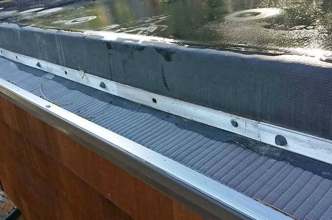 Close-up of a roof edge with a stainless steel micro-mesh gutter guard installed, preventing debris from entering. The wet roof and wood-paneled exterior are visible. Same-day pickup available at Home Depot.