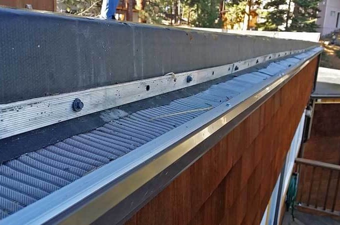 Close-up view of a house roof with a Gutter Guard by Gutterglove installed along the edge, designed to keep debris out of the gutter. Same-day pickup available. Trees and another building are visible in the background.