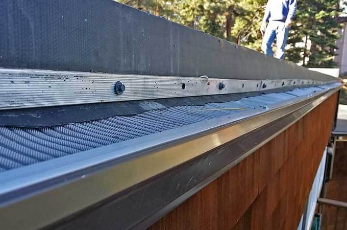 Close-up view of a Gutterglove gutter guard installed along the edge of a roof, with house siding below and trees in the background. A person stands on the roof in the upper right corner. Available at Home Depot with same-day pickup.