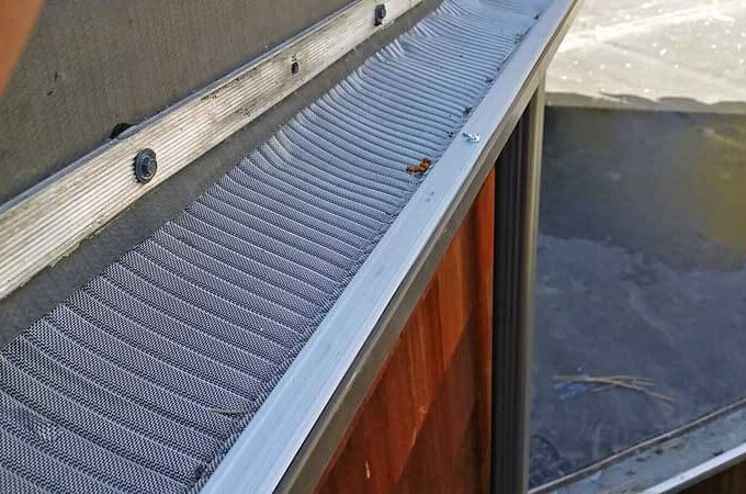 Close-up of a metal gutter guard featuring RiserStep Technology installed on the edge of a roof, with a few small bits of debris on top and a shingled roof visible in the background.