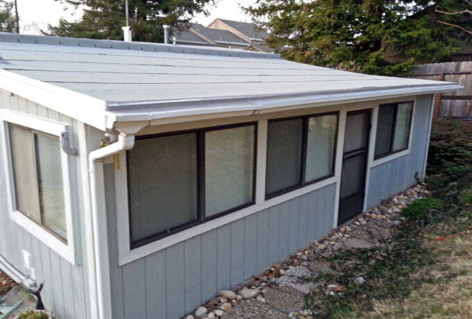 A small, light blue shed with a sloped metal roof features multiple windows with blinds, a black screen door, and a Gutter Guard by Gutterglove. Surrounded by rocks and grass, it sits in a backyard near trees and a wooden fence. Available at Home Depot.