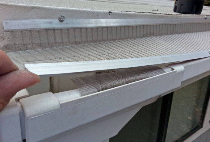 A close-up of a person installing a Gutterglove stainless steel micro-mesh gutter guard on the edge of a house roof to prevent debris from clogging the gutter. The person is holding a metal strip to secure the mesh in place.