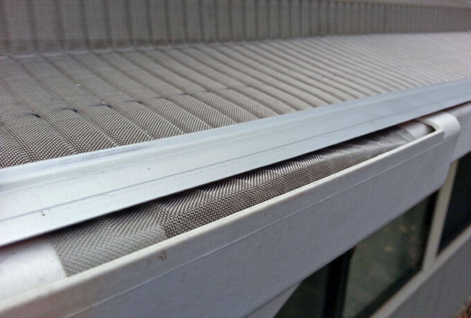 Close-up of a Home Depot stainless steel micro-mesh gutter guard installed along the edge of a roof, designed to prevent leaves and debris from clogging the gutter. The background shows part of an exterior wall and windows.