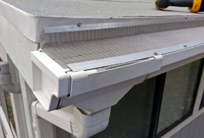 A close-up of a house roof corner shows white gutters with a stainless steel micro-mesh guard, a downspout, and a yellow-handled tool resting on the edge of the roof. Same-day pickup available.