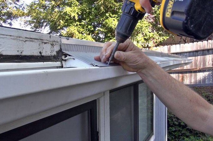 A person uses a power drill to install a metal Gutterglove guard over a rain gutter on the edge of a roof, with trees and a wooden fence in the background.
