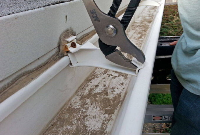 A person uses pliers to adjust a white plastic gutter bracket attached to a house while standing on a ladder. The dirty gutter, with visible debris inside, could benefit from a Gutterglove stainless steel micro-mesh found at Home Depot.