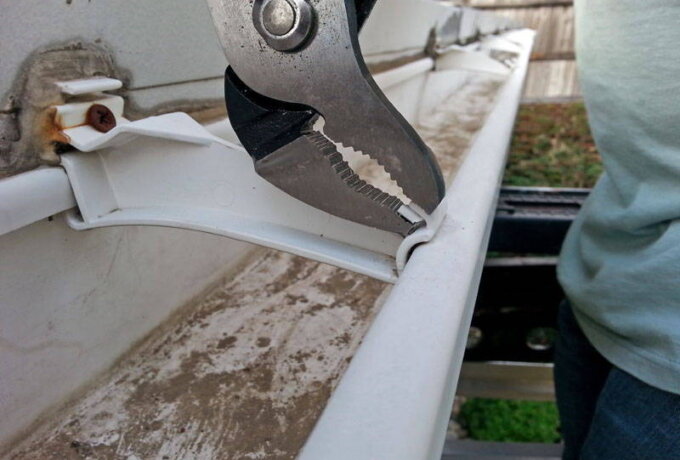 A pair of pliers is being used to repair or adjust a white plastic rain gutter attached to a building. The gutter appears dirty, and a person in a light shirt is standing nearby, possibly considering stainless steel micro-mesh for added protection.