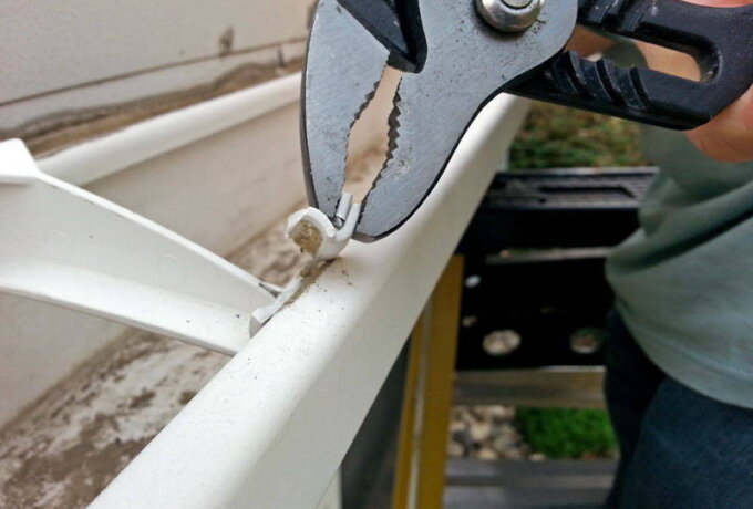A person uses pliers to remove a screw from a dirty white rain gutter attached to a house exterior, preparing for the installation of Gutter Guard by Gutterglove from Home Depot with same-day pickup.
