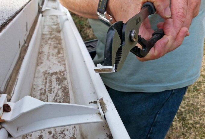 A person in a blue shirt uses pliers to remove a broken piece from a white rain gutter with dirt inside, ready for same-day pickup at Home Depot.