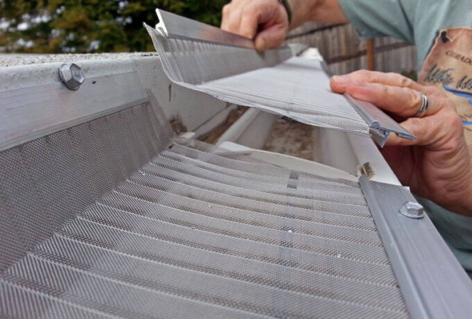 A person installs a Gutterglove mesh gutter guard on a house gutter, using their hands to fit the metal screen over the gutter to prevent debris from entering, utilizing RiserStep Technology for added protection.