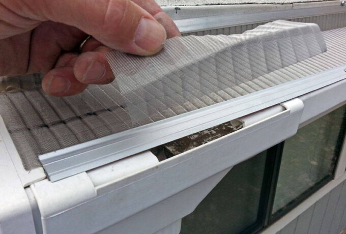 A hand lifts a section of Gutterglove stainless steel micro-mesh gutter guard from a white roof gutter, revealing the clean gutter underneath. The scene is outdoors with a window and downspout below.