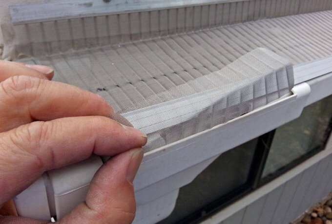 A person’s hand lifts a section of Gutter Guard by Gutterglove with RiserStep Technology from the edge of a white rain gutter attached to a house, revealing the underlying gutter and roof.