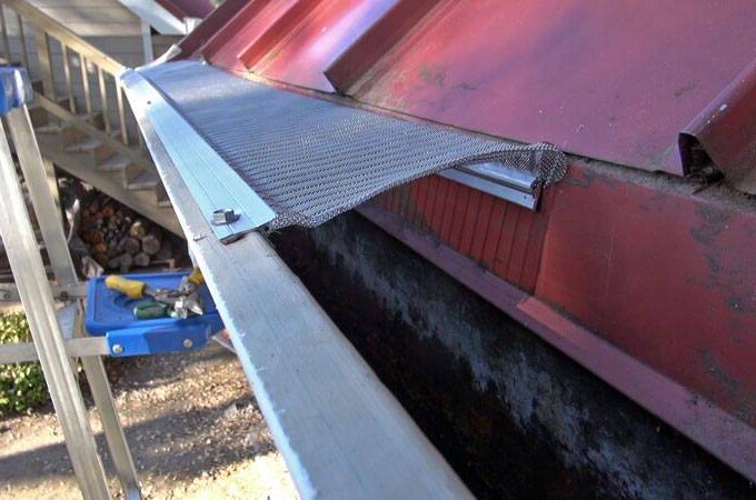 A close-up of a red metal roof with a mesh gutter guard partially installed. A ladder stands nearby, and tools are visible on a platform in the background—ideal for those seeking Gutterglove products with same-day pickup at Home Depot.