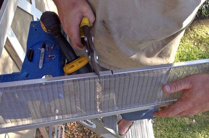A person uses tin snips to cut Gutter Guard by Gutterglove mesh while holding a drill, standing on a ladder next to a house. Tools are visible nearby, suggesting an easy DIY project with same-day pickup options at Home Depot.