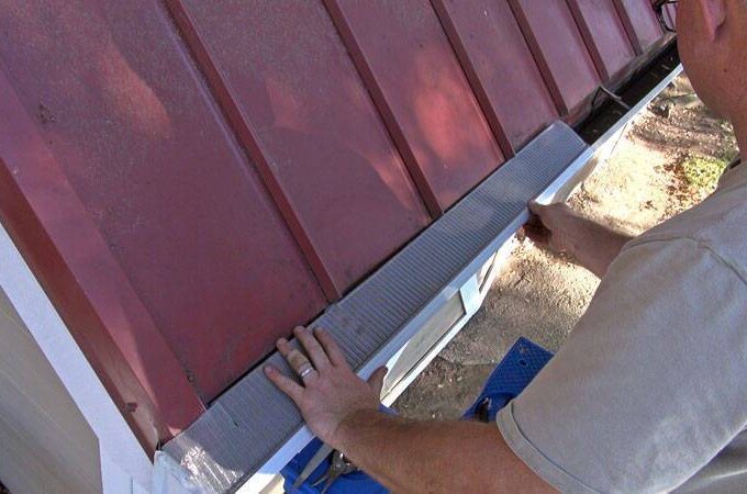 A person installs Gutter Guard by Gutterglove with RiserStep Technology along a metal roof edge to prevent debris from entering the gutter, standing on a ladder and pressing the guard into place.