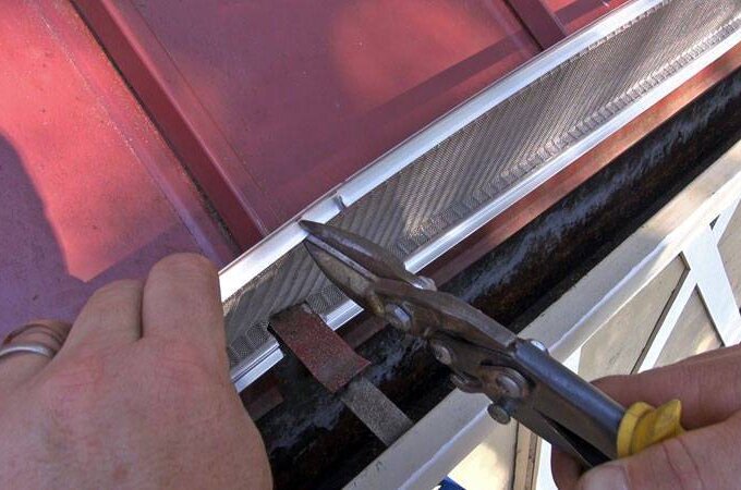 Close-up of hands using tin snips to cut a piece of metal screen or flashing along the edge of a red metal panel, possibly during a construction project. Get your materials fast with same-day pickup at Home Depot.